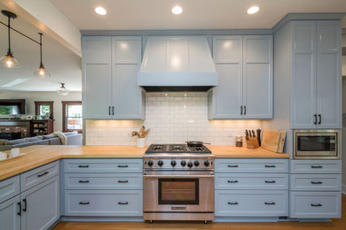 Large transitional galley light wood floor kitchen pantry photo in Portland with a farmhouse sink, recessed-panel cabinets, blue cabinets, wood countertops, white backsplash, subway tile backsplash, stainless steel appliances and a peninsula