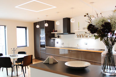 This is an example of a large modern single-wall open plan kitchen in London with an integrated sink, glass-front cabinets, grey cabinets, laminate countertops, white splashback, stainless steel appliances, medium hardwood flooring and an island.