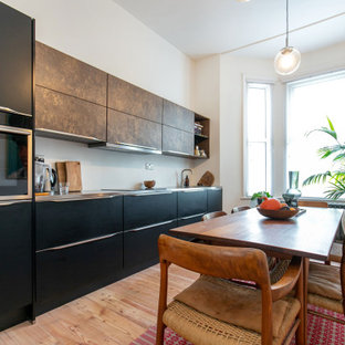 Bild på ett mellanstort funkis grå linjärt grått kök och matrum, med en integrerad diskho, luckor med upphöjd panel, svarta skåp, bänkskiva i rostfritt stål, vitt stänkskydd, stänkskydd i tegel, svarta vitvaror, ljust trägolv och beiget golv