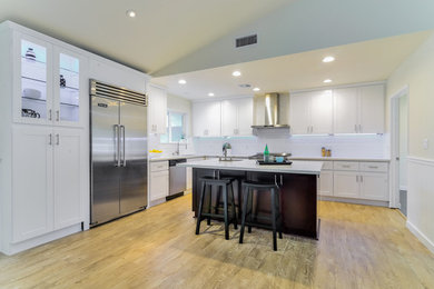 Example of a trendy l-shaped light wood floor kitchen design in Los Angeles with a farmhouse sink, white cabinets, white backsplash, subway tile backsplash, stainless steel appliances and an island