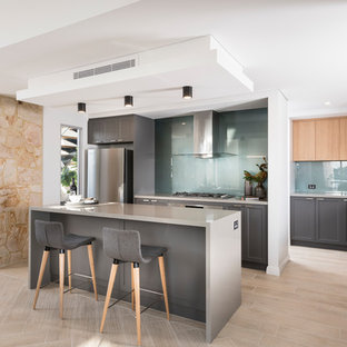 Ejemplo de cocina en L, actual, abierta, con puertas de armario grises, salpicadero azul, electrodomésticos de acero inoxidable, suelo de madera clara, una isla, suelo beige, armarios estilo shaker y salpicadero de vidrio templado