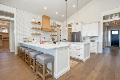 Photo of a large contemporary u-shaped open plan kitchen in Boise with a belfast sink, metro tiled splashback, stainless steel appliances, an island, brown floors, recessed-panel cabinets, white cabinets, composite countertops, white splashback, medium hardwood flooring and white worktops.