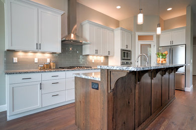 Example of a mid-sized transitional l-shaped dark wood floor and brown floor kitchen design in Portland with an undermount sink, shaker cabinets, white cabinets, granite countertops, gray backsplash, glass tile backsplash, stainless steel appliances, an island and multicolored countertops