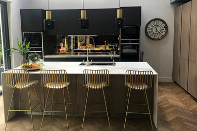 Photo of a contemporary open plan kitchen in Surrey with a submerged sink, flat-panel cabinets, black cabinets, composite countertops, metallic splashback, glass sheet splashback, black appliances, plywood flooring, an island, brown floors and white worktops.