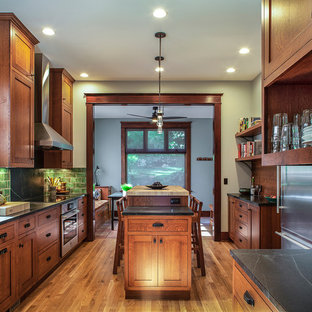 Large arts and crafts galley separate kitchen in Portland with a farmhouse sink, shaker cabinets, soapstone benchtops, green splashback, ceramic splashback, stainless steel appliances, medium hardwood floors, with island, medium wood cabinets, brown floor and black benchtop.