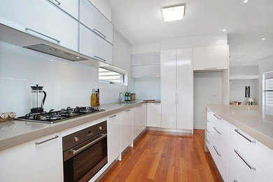 Cette image montre une cuisine ouverte linéaire design avec un placard à porte plane, des portes de placard blanches, une crédence blanche, un électroménager en acier inoxydable, un sol en bois brun et îlot.