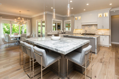 Large trendy u-shaped light wood floor and brown floor kitchen photo in Other with an undermount sink, shaker cabinets, white cabinets, marble countertops, beige backsplash, stone tile backsplash, stainless steel appliances, two islands and white countertops
