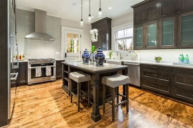 Example of a large u-shaped medium tone wood floor enclosed kitchen design in Seattle with a farmhouse sink, recessed-panel cabinets, gray cabinets, quartz countertops, white backsplash, ceramic backsplash, stainless steel appliances and an island