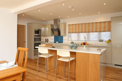 Inspiration for a medium sized contemporary l-shaped open plan kitchen in Kent with flat-panel cabinets, beige cabinets, blue splashback, glass sheet splashback, stainless steel appliances, medium hardwood flooring and an island.