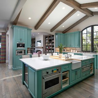 Idee per un'ampia cucina mediterranea con lavello stile country, ante con bugna sagomata, ante turchesi, paraspruzzi multicolore, elettrodomestici in acciaio inossidabile, pavimento in legno massello medio, isola, pavimento marrone e top bianco