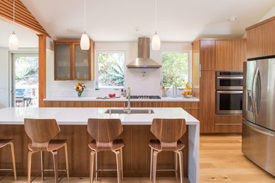 Modern inredning av ett stort kök, med en dubbel diskho, släta luckor, skåp i mellenmörkt trä, bänkskiva i kvarts, vitt stänkskydd, rostfria vitvaror, mellanmörkt trägolv och en köksö