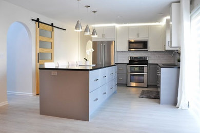 Example of a small minimalist l-shaped light wood floor and beige floor eat-in kitchen design in San Francisco with a drop-in sink, shaker cabinets, gray cabinets, quartz countertops, white backsplash, subway tile backsplash, stainless steel appliances, an island and black countertops