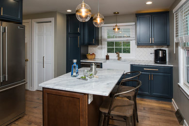 Example of a small country u-shaped medium tone wood floor enclosed kitchen design in New York with a farmhouse sink, recessed-panel cabinets, blue cabinets, marble countertops, white backsplash, subway tile backsplash, stainless steel appliances and an island