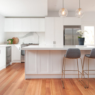 Large transitional l-shaped open plan kitchen in Melbourne with an undermount sink, white cabinets, white splashback, stone slab splashback, stainless steel appliances, medium hardwood floors, with island, white benchtop, shaker cabinets and beige floor.