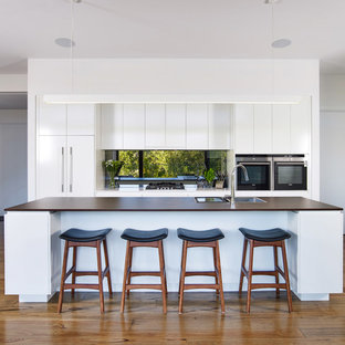 Inspiration for a large contemporary galley kitchen in Other with flat-panel cabinets, white cabinets, solid surface benchtops, metallic splashback, mirror splashback, black appliances, medium hardwood floors, with island, brown floor, brown benchtop and a drop-in sink.