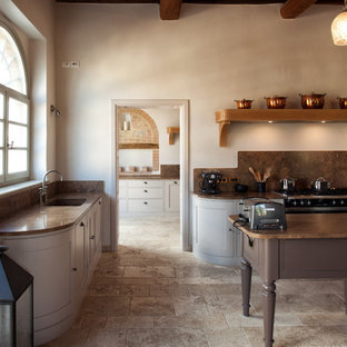 Imagen de cocina en L, campestre, abierta, con fregadero bajoencimera, armarios con rebordes decorativos, puertas de armario grises, encimera de mármol, salpicadero marrón, salpicadero de losas de piedra, electrodomésticos negros, suelo de piedra caliza y una isla