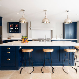 This is an example of a classic l-shaped kitchen in Cardiff with a belfast sink, shaker cabinets, blue cabinets, stainless steel appliances, an island, beige floors and white worktops.