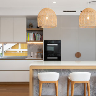 Large contemporary galley open plan kitchen in Brisbane with an undermount sink, shaker cabinets, white cabinets, solid surface benchtops, beige splashback, marble splashback, black appliances, medium hardwood floors, multiple islands, brown floor and white benchtop.