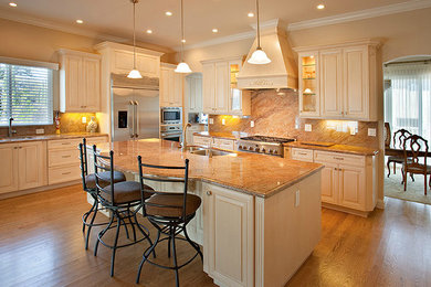 Inspiration for a large coastal l-shaped medium tone wood floor eat-in kitchen remodel in Other with a drop-in sink, white cabinets, stainless steel appliances, beige backsplash, an island, raised-panel cabinets, granite countertops and stone slab backsplash
