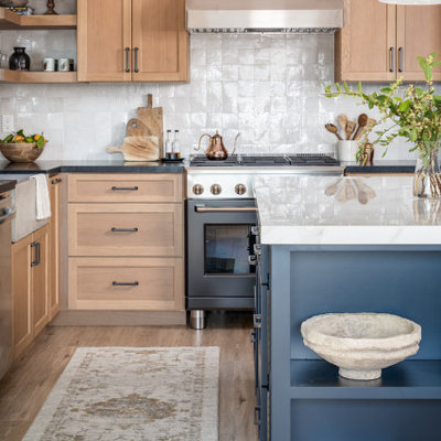 Example of a mid-sized transitional l-shaped vinyl floor and beige floor kitchen design in San Diego with a farmhouse sink, shaker cabinets, blue cabinets, solid surface countertops, white backsplash, terra-cotta backsplash, black appliances, an island and black countertops