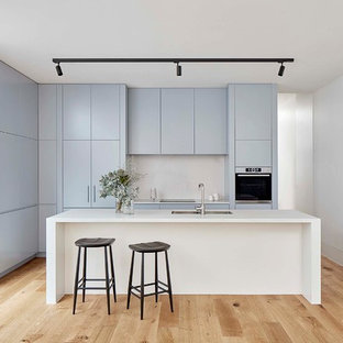 Inspiration for a mid-sized contemporary galley kitchen in Melbourne with a double-bowl sink, blue cabinets, quartz benchtops, white splashback, stone slab splashback, stainless steel appliances, light hardwood floors, with island, white benchtop, flat-panel cabinets and brown floor.