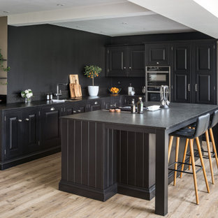 Photo of a large classic l-shaped open plan kitchen in Hampshire with raised-panel cabinets, black cabinets, an island, black worktops, a submerged sink, black appliances, medium hardwood flooring and brown floors.