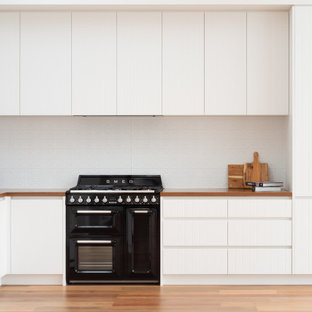 Small scandinavian l-shaped eat-in kitchen in Melbourne with a double-bowl sink, flat-panel cabinets, white cabinets, wood benchtops, white splashback, metal splashback, black appliances, medium hardwood floors, no island, brown floor, brown benchtop and vaulted.