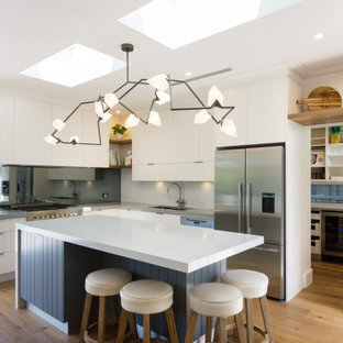 Beach style l-shaped open plan kitchen in Melbourne with an undermount sink, quartz benchtops, metallic splashback, stainless steel appliances, medium hardwood floors, with island, brown floor, flat-panel cabinets, white cabinets, mirror splashback and grey benchtop.