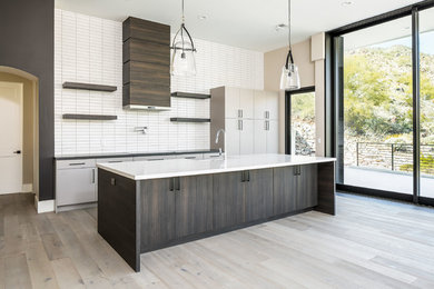 Large trendy l-shaped light wood floor and gray floor open concept kitchen photo in Phoenix with a farmhouse sink, flat-panel cabinets, quartz countertops, white backsplash, subway tile backsplash, stainless steel appliances, an island and white countertops