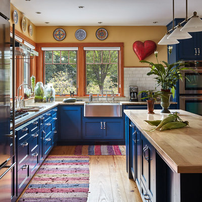 Example of a country l-shaped medium tone wood floor and brown floor kitchen design in Bridgeport with a farmhouse sink, shaker cabinets, blue cabinets, wood countertops, white backsplash, subway tile backsplash, stainless steel appliances, an island and brown countertops