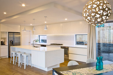 Large minimalist l-shaped light wood floor eat-in kitchen photo in Wellington with a double-bowl sink, flat-panel cabinets, white cabinets, solid surface countertops, stone slab backsplash, stainless steel appliances and an island