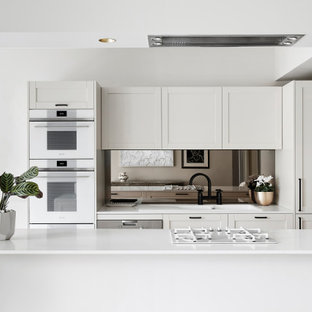 Mid-sized contemporary galley kitchen in Melbourne with a drop-in sink, shaker cabinets, white cabinets, mirror splashback, white appliances, with island and white benchtop.