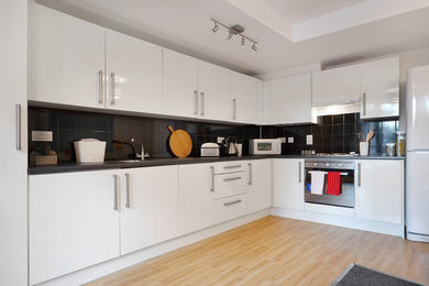 Photo of a contemporary kitchen in London with flat-panel cabinets, white cabinets, black splashback, stainless steel appliances and medium hardwood flooring.