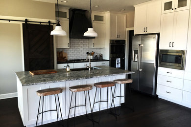 Example of a farmhouse l-shaped dark wood floor and brown floor eat-in kitchen design in Other with a farmhouse sink, raised-panel cabinets, white cabinets, granite countertops, white backsplash, subway tile backsplash, stainless steel appliances, an island and multicolored countertops
