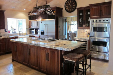 Large elegant l-shaped travertine floor enclosed kitchen photo in Los Angeles with a farmhouse sink, raised-panel cabinets, dark wood cabinets, granite countertops, white backsplash, ceramic backsplash, stainless steel appliances and an island