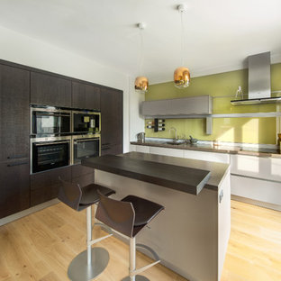 Inspiration for a small contemporary l-shaped kitchen/diner in Oxfordshire with an integrated sink, flat-panel cabinets, grey cabinets, composite countertops, green splashback, glass sheet splashback, black appliances, light hardwood flooring, an island and grey worktops.