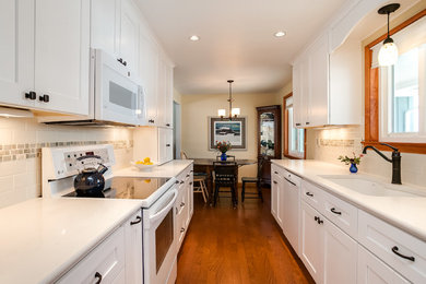 Example of a mid-sized classic galley medium tone wood floor eat-in kitchen design in Denver with an undermount sink, recessed-panel cabinets, white cabinets, solid surface countertops, white backsplash, ceramic backsplash and white appliances