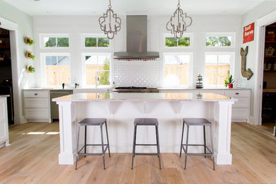 Open concept kitchen - mid-sized country l-shaped light wood floor open concept kitchen idea in Birmingham with a farmhouse sink, white backsplash, stainless steel appliances, an island, shaker cabinets, white cabinets, solid surface countertops and porcelain backsplash