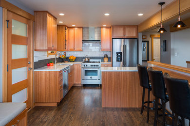 シアトルにあるコンテンポラリースタイルのおしゃれなキッチン (エプロンフロントシンク、シェーカースタイル扉のキャビネット、中間色木目調キャビネット、白いキッチンパネル、サブウェイタイルのキッチンパネル、シルバーの調理設備、濃色無垢フローリング、茶色い床、白いキッチンカウンター) の写真