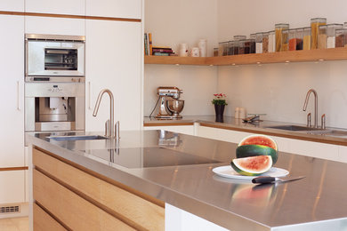 Idéer för att renovera ett mellanstort funkis grå grått kök med öppen planlösning, med en integrerad diskho, släta luckor, vita skåp, bänkskiva i rostfritt stål, glaspanel som stänkskydd, rostfria vitvaror och en köksö
