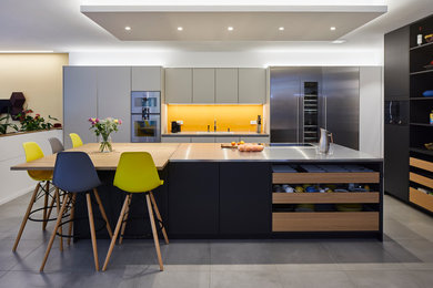 This is an example of a large modern single-wall open plan kitchen with black cabinets, stainless steel worktops, yellow splashback, glass sheet splashback, stainless steel appliances, ceramic flooring, an island and grey floors.