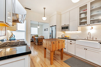 Example of a large cottage galley light wood floor and beige floor enclosed kitchen design in San Francisco with a farmhouse sink, shaker cabinets, white cabinets, granite countertops, white backsplash, subway tile backsplash, stainless steel appliances and an island