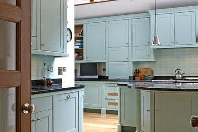 This is an example of a medium sized classic u-shaped open plan kitchen in London with a built-in sink, shaker cabinets, turquoise cabinets, granite worktops, black splashback, stone tiled splashback, black appliances, light hardwood flooring, an island, beige floors and black worktops.