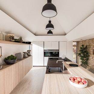 This is an example of a mid-sized contemporary single-wall open plan kitchen in Gloucestershire with an integrated sink, flat-panel cabinets, white cabinets, solid surface benchtops, black appliances, light hardwood floors, a peninsula, beige floor and brown benchtop.