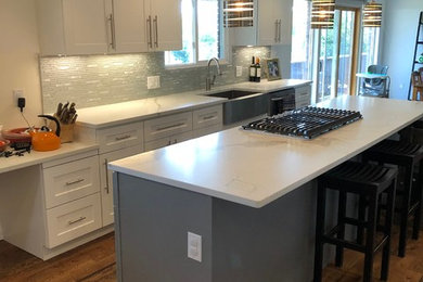 Example of a mid-sized transitional galley dark wood floor and brown floor eat-in kitchen design in Denver with a farmhouse sink, shaker cabinets, white cabinets, gray backsplash, stainless steel appliances, an island and white countertops