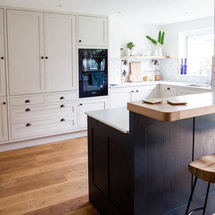 This is an example of a medium sized contemporary u-shaped kitchen in Other with a double-bowl sink, shaker cabinets, beige cabinets, white splashback, ceramic splashback, stainless steel appliances, light hardwood flooring, a breakfast bar, brown floors and white worktops.