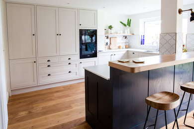 This is an example of a medium sized contemporary u-shaped kitchen in Other with a double-bowl sink, shaker cabinets, beige cabinets, white splashback, ceramic splashback, stainless steel appliances, light hardwood flooring, a breakfast bar, brown floors and white worktops.