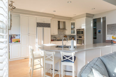 Example of a classic light wood floor kitchen design in Vancouver with an undermount sink, raised-panel cabinets, white cabinets, quartz countertops, gray backsplash, stone tile backsplash, stainless steel appliances and a peninsula