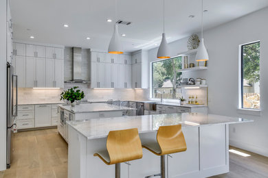 Photo of a large contemporary u-shaped open plan kitchen in Austin with shaker cabinets, white cabinets, white splashback, light hardwood flooring, multiple islands, beige floors, a belfast sink, quartz worktops, stone tiled splashback, stainless steel appliances and white worktops.