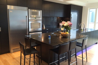 This is an example of a large contemporary single-wall kitchen/diner in Calgary with a submerged sink, flat-panel cabinets, black cabinets, quartz worktops, black splashback, stainless steel appliances, light hardwood flooring and an island.